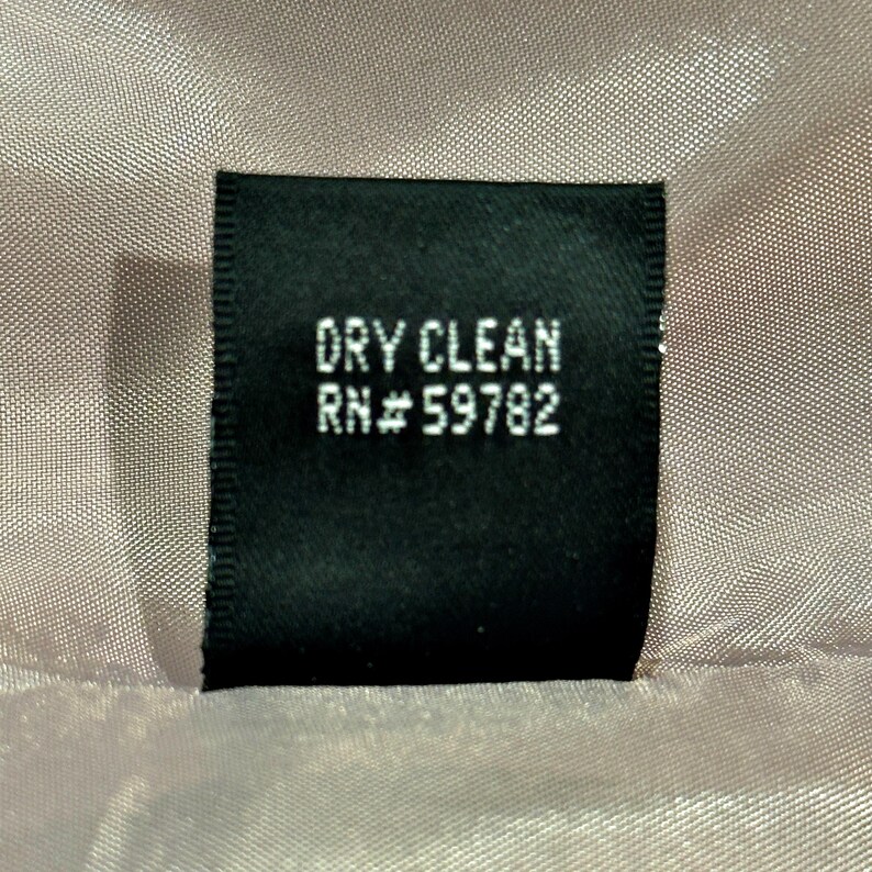 K&ouml;nnte beinhalten: Ein schwarzes Etikett mit dem wei&szlig;en Aufdruck "DRY CLEAN" und "RN# 59782". Das Etikett befindet sich vor einem strukturierten, hellrosa Stoff.