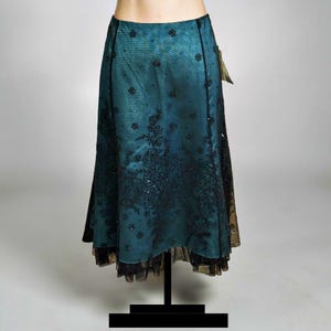 Op de afbeelding: Een teal rok met zwarte bloemenborduursels en pailletten. De rok heeft een gelaagd ontwerp met een zwarte kanten zoom en wordt gepresenteerd op een zwarte mannequinstandaard. Er is een label aan de rok bevestigd.