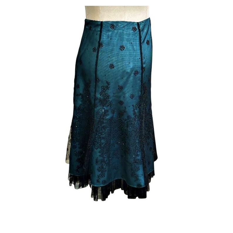 Op de afbeelding: Een teal rok met zwarte kanten details. De rok heeft een getailleerde taille en een uitlopende zoom, met ingewikkelde bloempatronen en een zwarte kanten rand. De rok is gemaakt van een glanzende stof en heeft een gelaagd ontwerp.