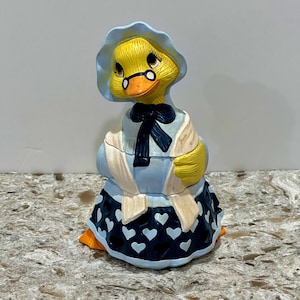 May include: A ceramic duck figurine, painted in yellow, blue, and white, with a bonnet and a bow. The duck is wearing a blue dress with white heart patterns. The figurine has orange feet and a white shawl.