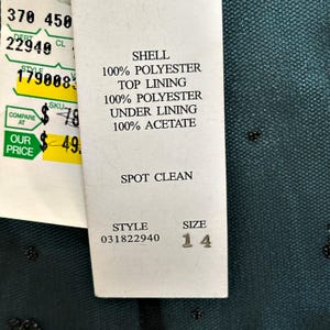 Op de afbeelding: Een kledinglabel met details over de stof samenstelling. Het label vermeldt "SHELL 100% POLYESTER", "TOP LINING 100% POLYESTER" en "UNDER LINING 100% ACETATE". De maat is 14. Het artikelnummer is 031822940. Het label zegt ook "SPOT CLEAN".