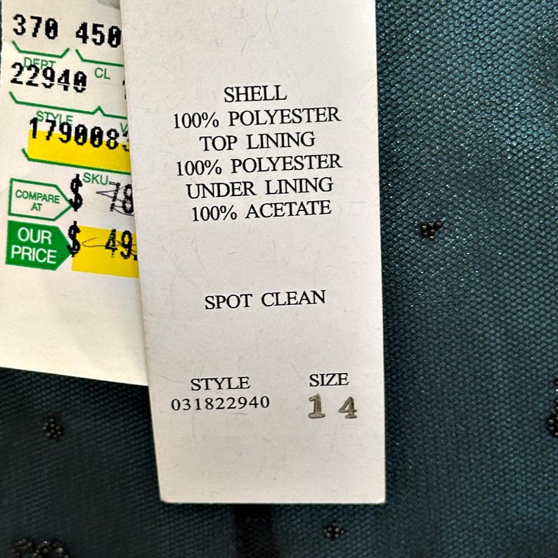Op de afbeelding: Een kledinglabel met details over de stof samenstelling. Het label vermeldt "SHELL 100% POLYESTER", "TOP LINING 100% POLYESTER" en "UNDER LINING 100% ACETATE". De maat is 14. Het artikelnummer is 031822940. Het label zegt ook "SPOT CLEAN".