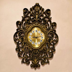 Könnte beinhalten: Antike Wanduhr mit goldfarbenem Rahmen und Zifferblatt. Die Uhr hat römische Ziffern und dekorative Schnörkel. Das Zifferblatt der Uhr ist hellgoldfarben, der Rahmen ist ein dunkleres, gealtertes Gold.