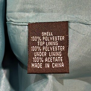 Op de afbeelding: Close-up van een bruin stoffen label met witte tekst. Het label luidt "SHELL 100% POLYESTER TOP LINING 100% POLYESTER UNDER LINING 100% ACETATE MADE IN CHINA". Het label is in een turquoise stof genaaid.