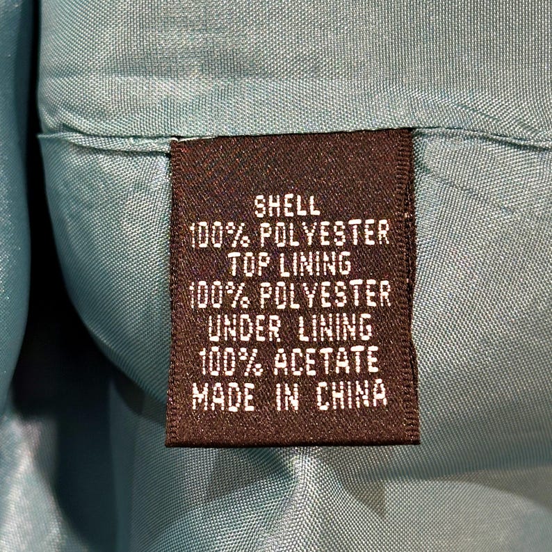 Op de afbeelding: Close-up van een bruin stoffen label met witte tekst. Het label luidt "SHELL 100% POLYESTER TOP LINING 100% POLYESTER UNDER LINING 100% ACETATE MADE IN CHINA". Het label is in een turquoise stof genaaid.