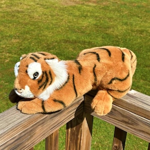 May include: A plush tiger toy in shades of orange, brown, black, and white, resting on a wooden railing. The toy has black stripes, a white collar, and detailed facial features.