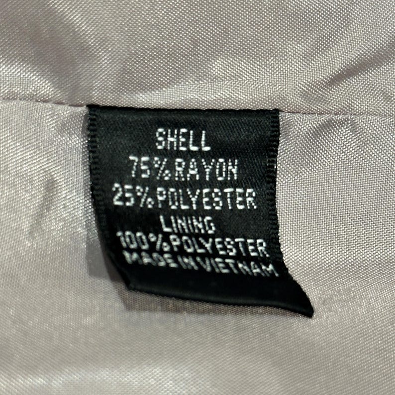 K&ouml;nnte beinhalten: Nahaufnahme eines schwarzen Kleidungsetiketts mit wei&szlig;em Text. Das Etikett lautet "SHELL 75% RAYON 25% POLYESTER LINING 100% POLYESTER MADE IN VIETNAM". Das Etikett ist in einen hellbeigen Stoff eingen&auml;ht.