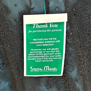 Op de afbeelding: Een teal kledingstuk met ingewikkelde kralen en een label met de tekst "Thank You for purchasing this garment." Het label bevat ook informatie over retourneren en ruilen, en het Stein Mart-logo.