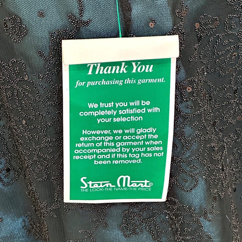 Op de afbeelding: Een teal kledingstuk met ingewikkelde kralen en een label met de tekst "Thank You for purchasing this garment." Het label bevat ook informatie over retourneren en ruilen, en het Stein Mart-logo.