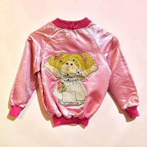 May include: A light pink satin bomber jacket featuring a ribbed collar, cuffs, and hem. The back displays a large embroidered applique of a doll with yellow yarn hair, wearing a white dress with floral accents and pink hair ribbons.