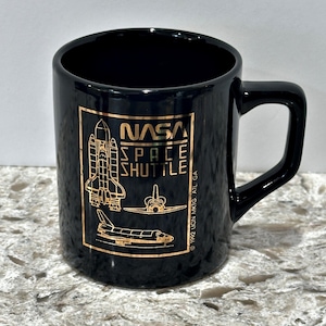 Könnte beinhalten: Schwarze NASA Space Shuttle-Tasse mit goldfarbenen Grafiken. Die Tasse zeigt das NASA-Logo und Illustrationen von Raumfähren. Der Text "NASA Space Shuttle" ist deutlich sichtbar. Die Tasse hat einen Henkel und eine glänzende Oberfläche.