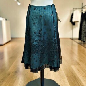 Op de afbeelding: Teal rok met zwarte bloemenborduursels en pailletten accenten. De rok heeft een gelaagd ontwerp met een geplooid zoom en wordt gepresenteerd op een mannequin. De rok is A-lijn en heeft een lichte glans.