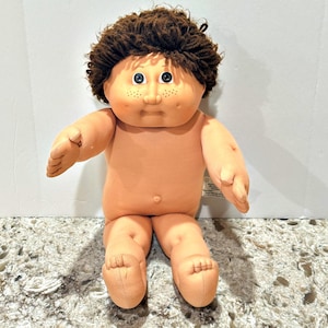 May include: A doll with a soft, light-toned fabric body and a round, vinyl head featuring dark brown yarn hair, blue eyes, and freckles across the cheeks. The doll has a neutral expression, a small belly button, and is posed in a seated position.
