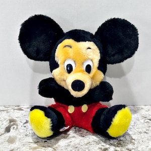 May include: A plush toy featuring a black and tan character with large round ears, white eyes with black pupils, a black nose, and red pants with two yellow oval buttons. The character has yellow feet and is depicted in a seated position.