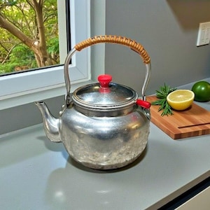 May include: A vintage silver tea kettle with a red knob and handle accents. The handle is wrapped in a woven material. A wooden chopping board with a lemon and lime sits nearby, suggesting a kitchen setting.