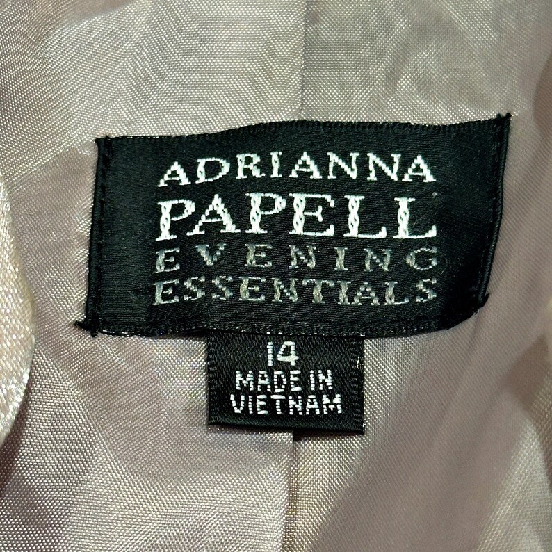 K&ouml;nnte beinhalten: Nahaufnahme eines schwarzen Etiketts mit wei&szlig;em Text, der "ADRIANNA PAPELL EVENING ESSENTIALS" lautet. Darunter befindet sich ein kleineres Etikett mit der Aufschrift "14 MADE IN VIETNAM".