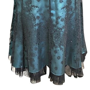 Op de afbeelding: Een teal-kleurige jurk met zwarte kanten overlay en pailletten accenten. De jurk heeft een gelaagde rok met een geschulpte zoom. De kant cre&euml;ert een bloemenpatroon en voegt elegantie toe.