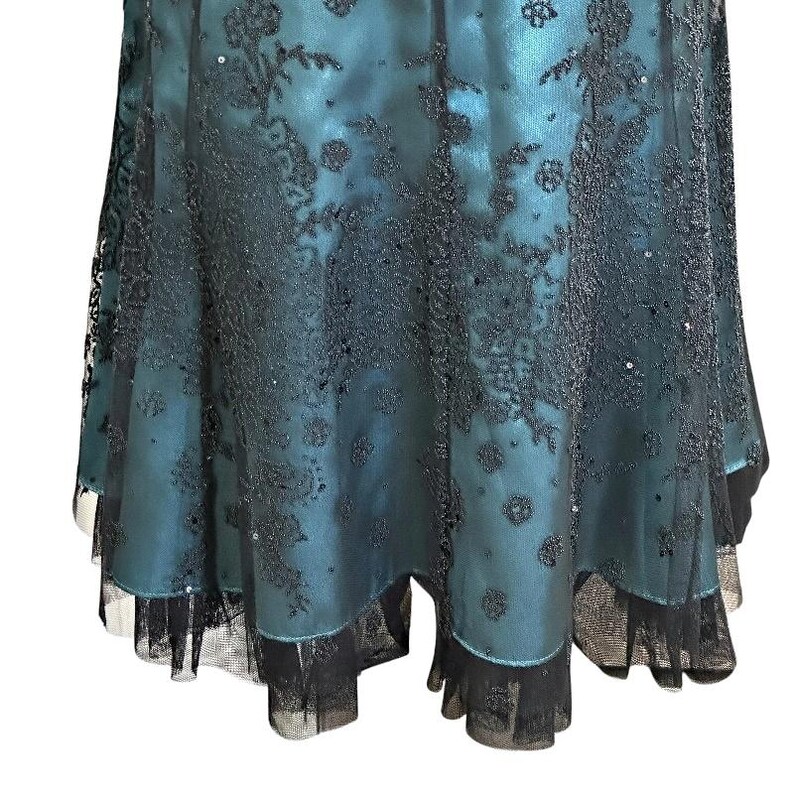 Op de afbeelding: Een teal-kleurige jurk met zwarte kanten overlay en pailletten accenten. De jurk heeft een gelaagde rok met een geschulpte zoom. De kant cre&euml;ert een bloemenpatroon en voegt elegantie toe.