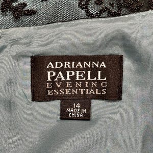 Op de afbeelding: Close-up van een zwart label met witte tekst "ADRIANNA PAPELL EVENING ESSENTIALS". Onder de tekst staat een kleiner label met de tekst "14 MADE IN CHINA". Het label staat op een teal stof.