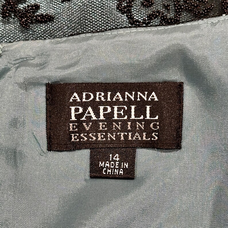 Op de afbeelding: Close-up van een zwart label met witte tekst "ADRIANNA PAPELL EVENING ESSENTIALS". Onder de tekst staat een kleiner label met de tekst "14 MADE IN CHINA". Het label staat op een teal stof.