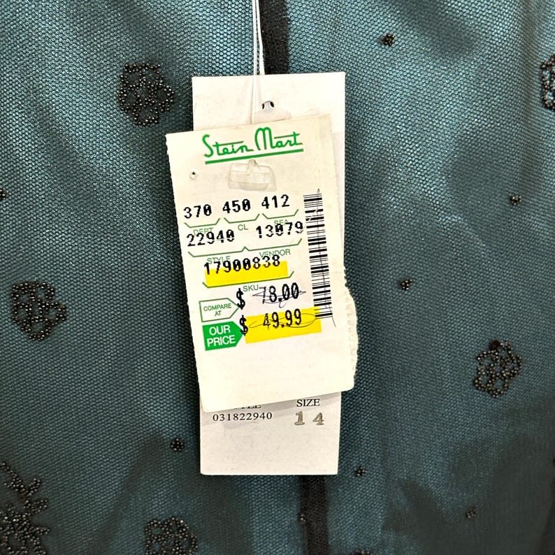 Op de afbeelding: Een teal-kleurig kledingstuk met zwarte kralenversieringen. Een wit label van Stein Mart is bevestigd, met productdetails en maat 14. Het label bevat de tekst "OUR PRICE $49.99".