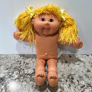 Puede incluir: Una muñeca con cuerpo de tela color canela suave y cabeza de vinilo con cabello de lana amarillo brillante peinado en dos coletas con lazos amarillos. La muñeca tiene mejillas redondas, ojos oscuros, boca pequeña, ombligo y dedos en los pies.