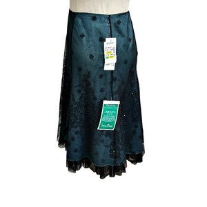 Op de afbeelding: Een teal-kleurige rok met een zwarte kanten overlay en een gezoomde zoom. De rok heeft een bloemenpatroon en een label met tekst. De rok wordt op een mannequin gepresenteerd.