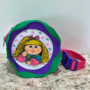 Könnte beinhalten: Grüne, lila und rosa Cabbage Patch Kids Bauchtasche. Die runde Tasche hat eine Cabbage Patch Kids Puppen-Grafik auf der Vorderseite. Der verstellbare Gurt ist rosa mit einer lila Schnalle. Die Tasche hat einen Reißverschluss.