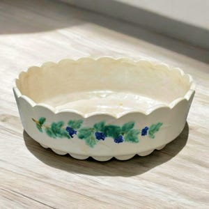 Puede incluir: Cuenco de cerámica color crema con borde festoneado. El cuenco está decorado con un diseño pintado a mano de hojas verdes y bayas azules. El cuenco está sobre una superficie de madera.