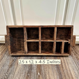 Puede incluir: Una caja de almacenamiento rectangular de madera marrón oscuro con ocho compartimentos internos y asas laterales. La caja presenta un acabado desgastado y veta de madera visible. El texto 25 x 12 x 4.5 Inches aparece impreso debajo.