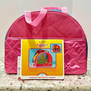 Może przedstawiać: Różowa torba wystawowa Hallmark Rainbow Brite z pasującym różowym uchwytem i przednią kieszenią. Torba znajduje się obok pudełka z napisem "Rainbow Land Display Carrier" i zdjęciem torby z postaciami Rainbow Brite.