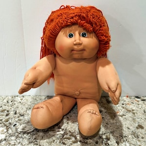 Könnte beinhalten: Eine weiche Puppe mit heller Haut, einem runden Gesicht und grünen Augen. Die Puppe hat einen ausgeprägten Bauchnabel und dickes, strukturiertes orangefarbenes Garnhaar, das wie eine Mütze geformt ist. Sie hat rosige Wangen und einen neutralen Ausdruck.