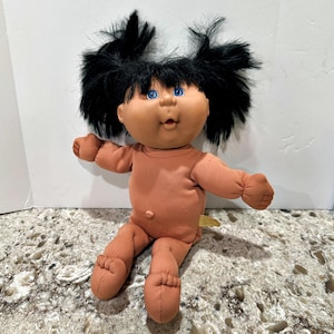 Peut inclure: Une poupée avec un corps en tissu beige doux, une tête ronde en vinyle et des yeux bleu vif. La poupée présente des cheveux noirs ébouriffés coiffés en deux couettes hautes, un petit nez et une bouche ouverte. Les membres sont arrondis.
