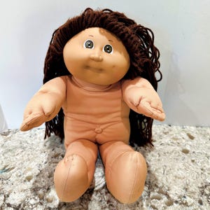 May include: A soft-bodied doll with a round, peach-toned fabric face, dimpled cheeks, and a small nose. The doll features dark brown yarn hair styled in long strands and large, round eyes with brown irises. It has a simple, unadorned fabric torso.