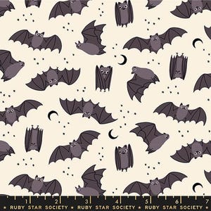 May include: A cream-coloured fabric featuring a repeating pattern of dark purple cartoon bats in various flight positions and hanging upside down, interspersed with small black stars and crescent moons. A ruler shows 18 cm at the bottom.