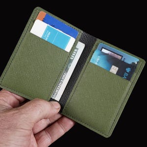 May include: Olive green bi-fold wallet with multiple card slots and a cash compartment. The wallet is open, revealing credit cards and a US one hundred dollar bill. The wallet is held against a black background.