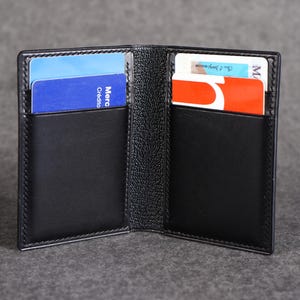 May include: A black leather card holder with a textured exterior and smooth interior pockets. The open wallet features two vertical slots on each side, currently holding four assorted credit and identification cards with visible text and colors.