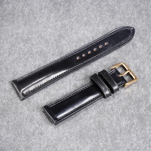 May include: A black leather watch strap with gold-colored buckle and hardware. The strap is made of shiny, smooth leather and is shown on a gray background. The strap is approximately 9 inches long.