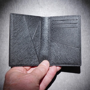 May include: Open black leather wallet with multiple card slots and a textured surface. The wallet is held open, showcasing its interior design and construction. The wallet is a dark gray color.