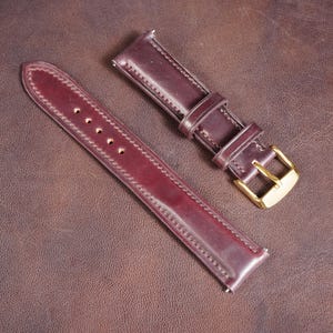 May include: A two-piece burgundy leather watch strap featuring a smooth finish, tonal stitching along the edges, and a polished gold-tone metal buckle. The strap includes a series of punched holes for adjustment and two leather keepers.
