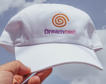 May include: A white baseball cap featuring an embroidered orange spiral logo above the purple text Dreamcast. The cap has a structured crown and a curved brim, showcasing the iconic branding clearly on the front panel of the headwear.