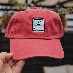 May include: A red baseball cap featuring an embroidered pixel art graphic of a vintage computer screen with a smiling face. The cap has a curved brim, a structured front panel, and stitched eyelets on the crown for ventilation and durability.