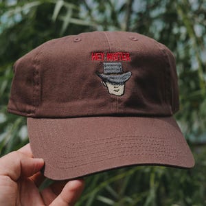 Op de afbeelding: Bruine baseballcap met de geborduurde tekst "HEY MISTER" in het rood. De cap heeft een ontwerp van een man met een hoed. Een klassiek, casual accessoire.