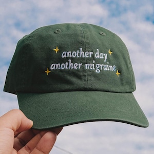 Op de afbeelding: Een donker olijfgroene baseballpet met wit geborduurde tekst "another day another migraine" en kleine gele vierpuntige sterren die rondom de tekst op het voorpaneel van de pet zijn geplaatst.