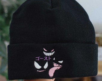 May include: A black knit beanie featuring an embroidered design of three stylized ghost-like faces in white and pink, with the Japanese text "ゴースト" in pink thread positioned above the lower left face on the folded cuff.