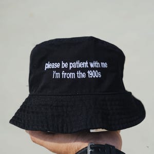 Peut inclure: Chapeau bob noir avec inscription brod&eacute;e en blanc "please be patient with me i'm from the 1900s"