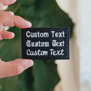 May include: A black rectangular patch with white embroidered text. The text reads "Custom Text" in three lines, with the middle line in a decorative font. The patch is held up by a hand.