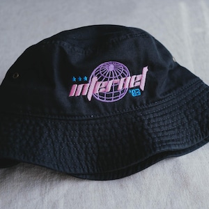 May include: Black bucket hat with embroidered "Infernel" text and globe graphic in pink and purple. The hat also features the number "83" in blue. A casual, stylish accessory.