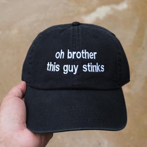 Peut inclure: Casquette de baseball noire avec l'inscription blanche brodée "oh brother this guy stinks". La casquette a une visière incurvée et une fermeture à sangle en tissu. Un accessoire décontracté.