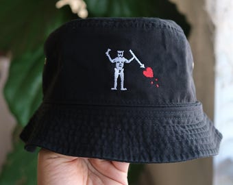 Könnte beinhalten: Ein schwarzer Fischerhut mit einer Stickerei, die eine hellgraue skelettartige Figur zeigt, die einen Speer auf ein rotes Herz richtet, wobei kleine rote Tropfen unter dem Herz auf der Vorderseite des Hutes zu sehen sind.